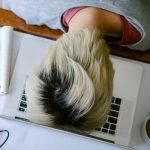 From above of tired fatigued unrecognizable female in casual wear resting head on netbook keyboard near notebook and cup of tea after long working day