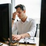 A tired call center employee with headset looks stressed at his office desk.